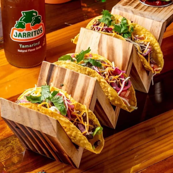 Four Hermanos Cantina Grill tacos with meat, cheese, onions, and cilantro in wooden holders, next to a Jarritos soda.
