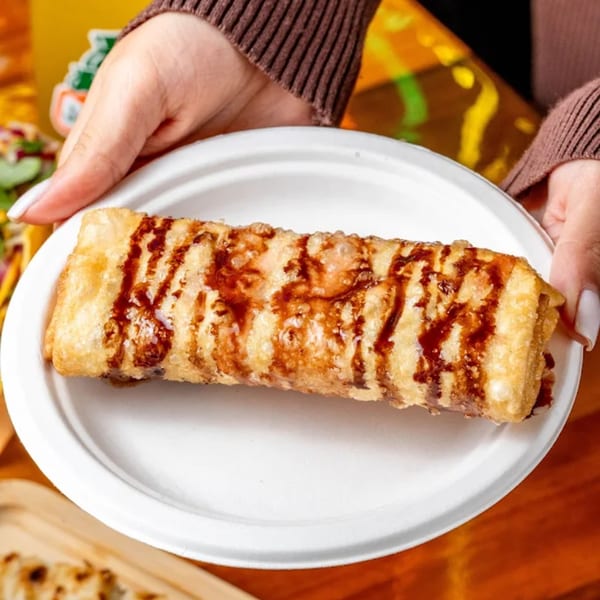 Hands hold a white plate with a golden-brown, fried dessert drizzled with chocolate sauce.