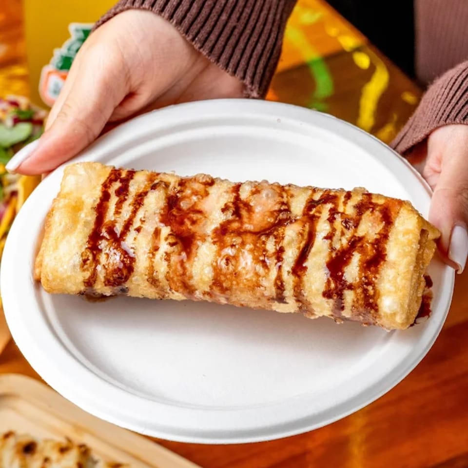 Hands hold a white plate with a golden-brown, fried dessert drizzled with chocolate sauce.