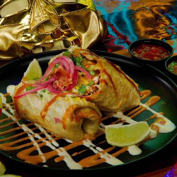 Two golden-brown burritos drizzled with white and orange sauces, topped with pickled onions and lime wedges.