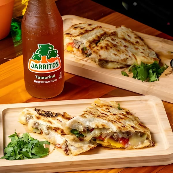 Two cheesy quesadillas with meat and vegetables on wooden boards, with a Jarritos Tamarind soda.